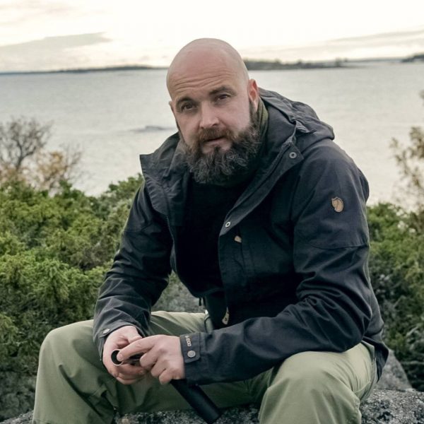Ukko Finlandin Jani Jaatinen istuu kivellä pidellen Ukko Snaps tuotetta käsissään yllään erähenkiset ulkoiluvaatteet. Taustalla näkyy merimaisema ja katajapuskia.