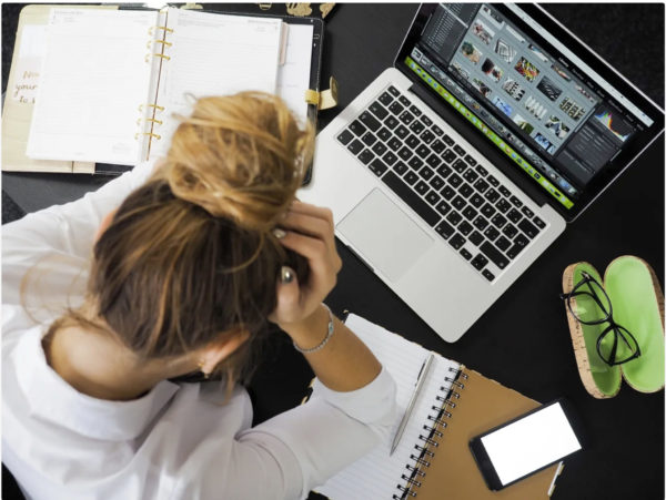 Stressaantuneen oloinen nainen istuu kannettavan tietokoneen ääressä ja haroo hiuksiaan. Pöydällä on avonaisia kirjoituslehtiöitä ja kännykkä.
